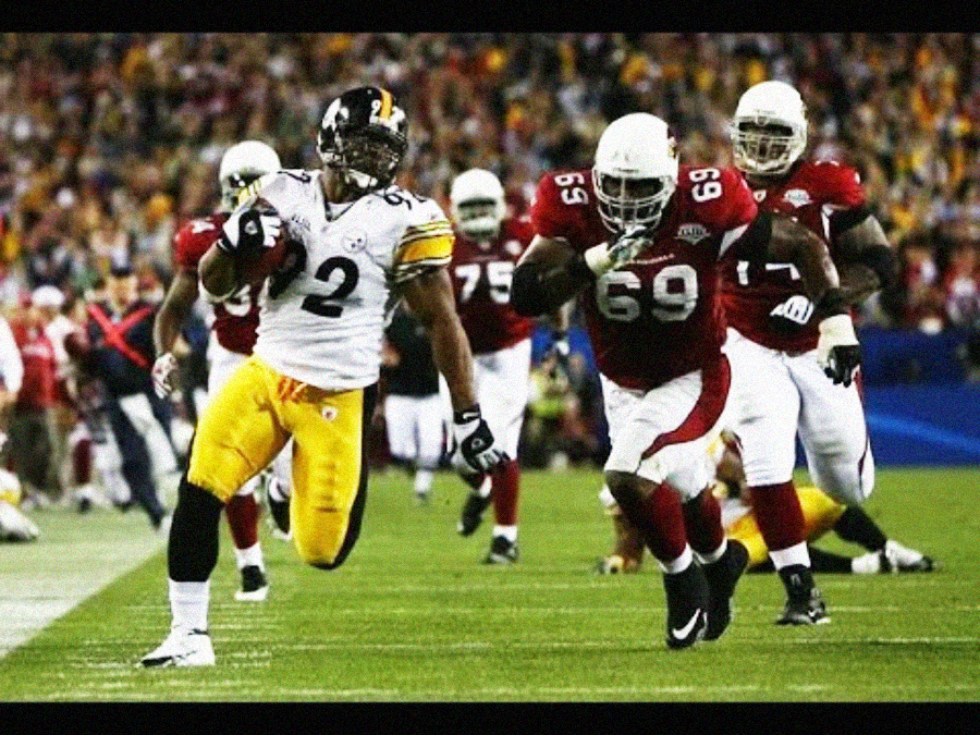 A football player in a yellow and black uniform runs alongside a player in a red and white uniform holding the ball, during a game with a crowd in the background. Other players are visible further behind on the field.