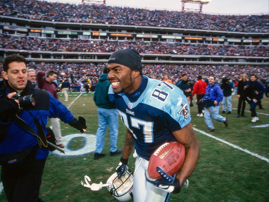 A smiling football player in a blue Tennessee Titans jersey, number 87, holds a helmet and football while running on the field. Photographers and a large cheering crowd fill the stadium in the background.
