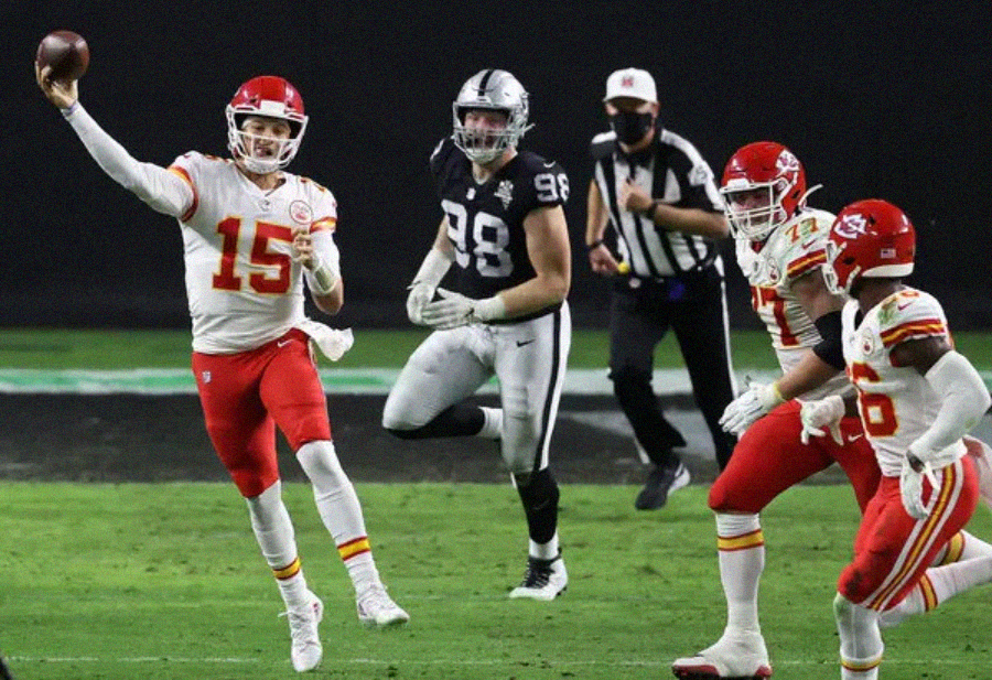 A football player in a red Kansas City Chiefs uniform throws a pass while being pursued by a Las Vegas Raiders defender. Two teammates block and an official watches in the background during a game.