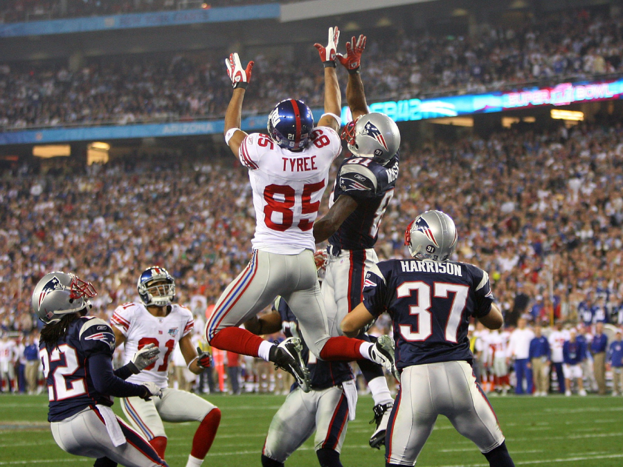 A New York Giants player jumps to catch a football, surrounded by New England Patriots defenders, during an NFL game in a crowded stadium.
