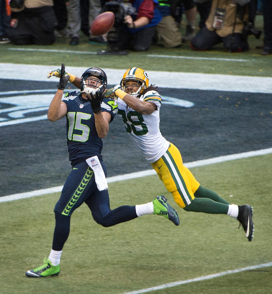 A Seattle Seahawks player reaches to catch a football in the end zone while a Green Bay Packers defender stretches to block the pass during an NFL game.