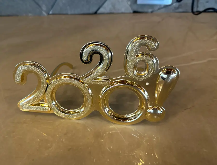 Gold novelty glasses shaped like the numbers "2026!" sit on a tan marble surface, with the numbers forming the frames for the lenses.