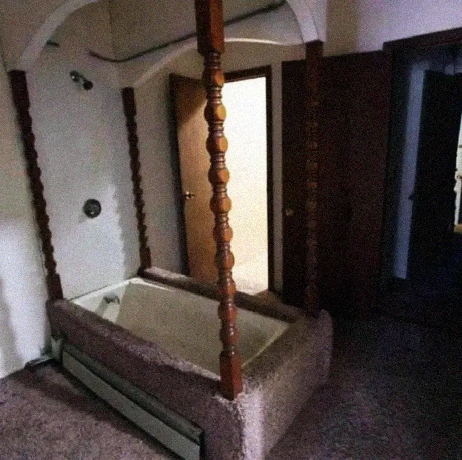 A bathtub surrounded by carpeted flooring and wooden columns, set in a corner of a dimly lit room with open doors and arched architectural details.
