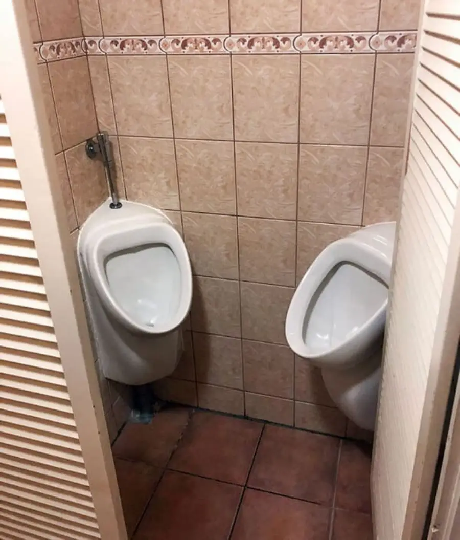 Two urinals are installed very close together in a small tiled restroom with no divider between them, making the space awkward and lacking privacy.