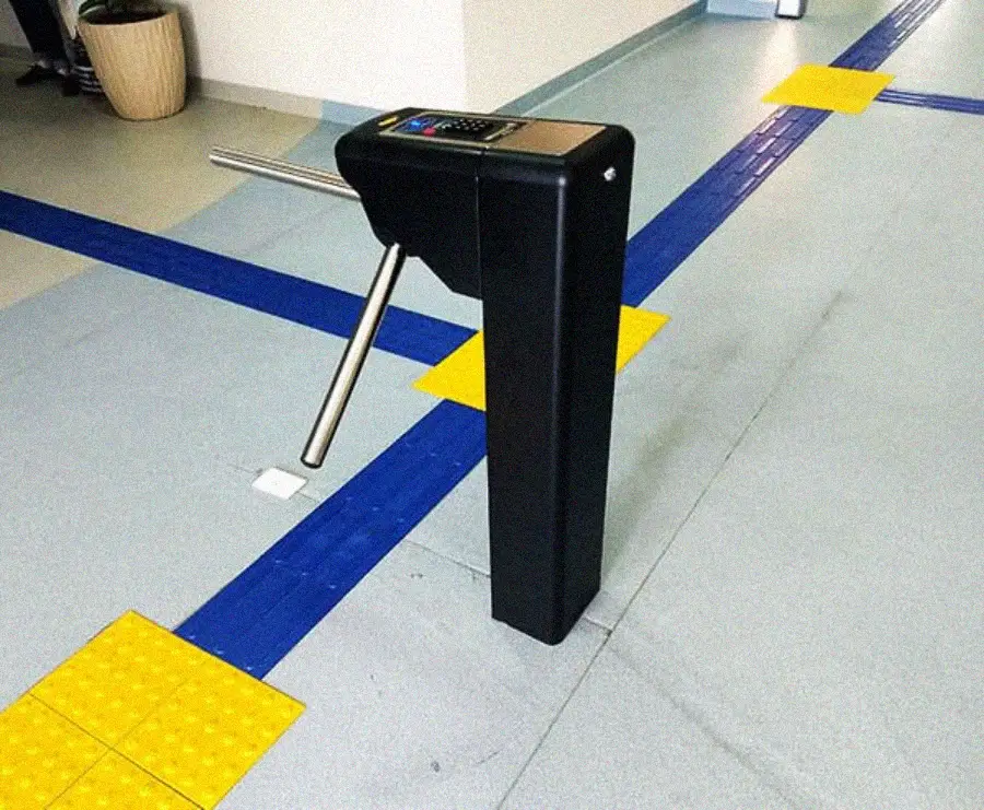 A black waist-high turnstile with two horizontal arms is installed in a hallway with blue tactile paving strips and yellow warning tiles on a light gray floor. A potted plant is in the corner.