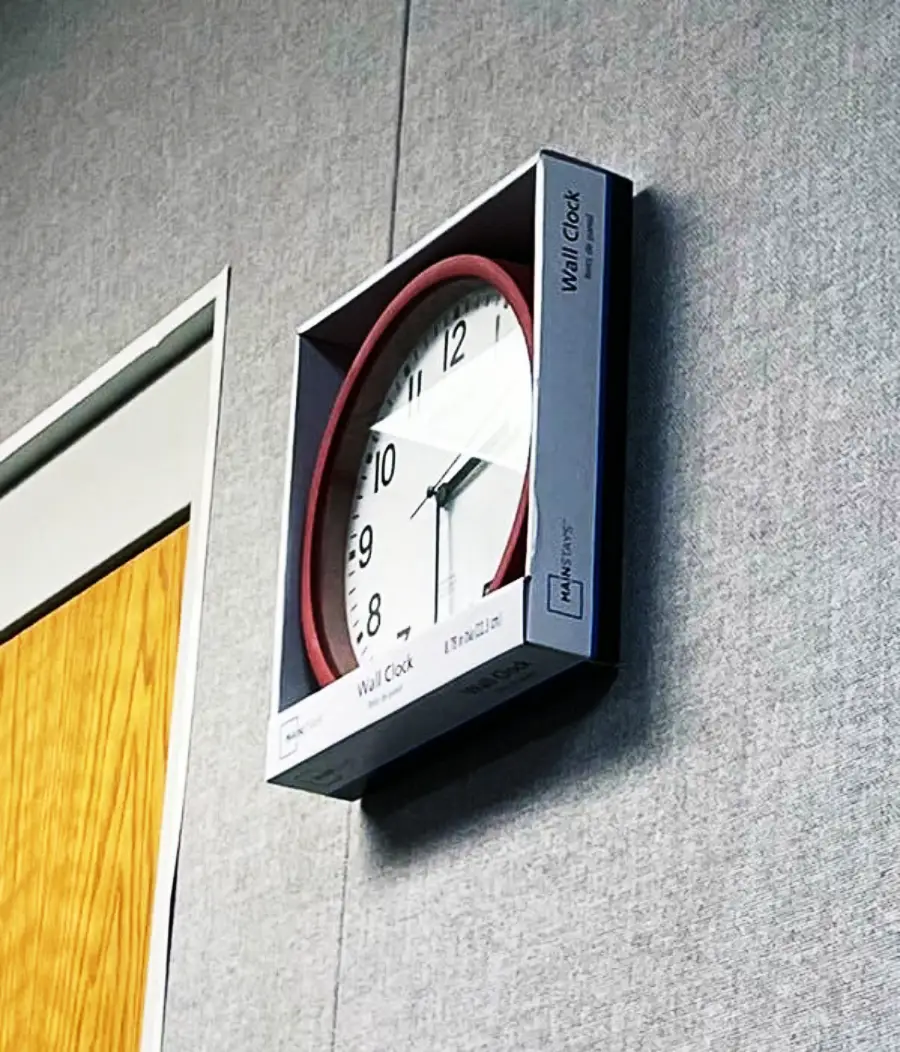 A wall clock is hanging on a gray wall, still inside its original square packaging. The box surrounds the clock, which shows the time as 11:54. A wooden door is visible to the left.