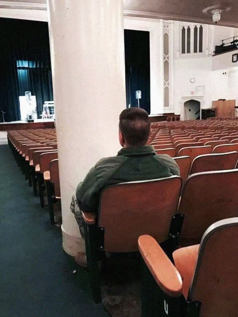 A person sits alone in an empty auditorium, facing a stage with blue curtains. Rows of vacant wooden seats surround them, and a large white pillar partially blocks the view.