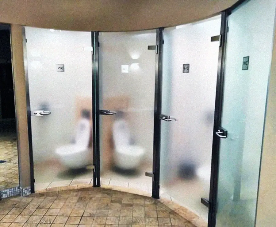 Three bathroom stalls with frosted glass doors provide only partial privacy, as the blurred doors still allow the toilets inside each stall to be visible from the outside. The floor is tiled.