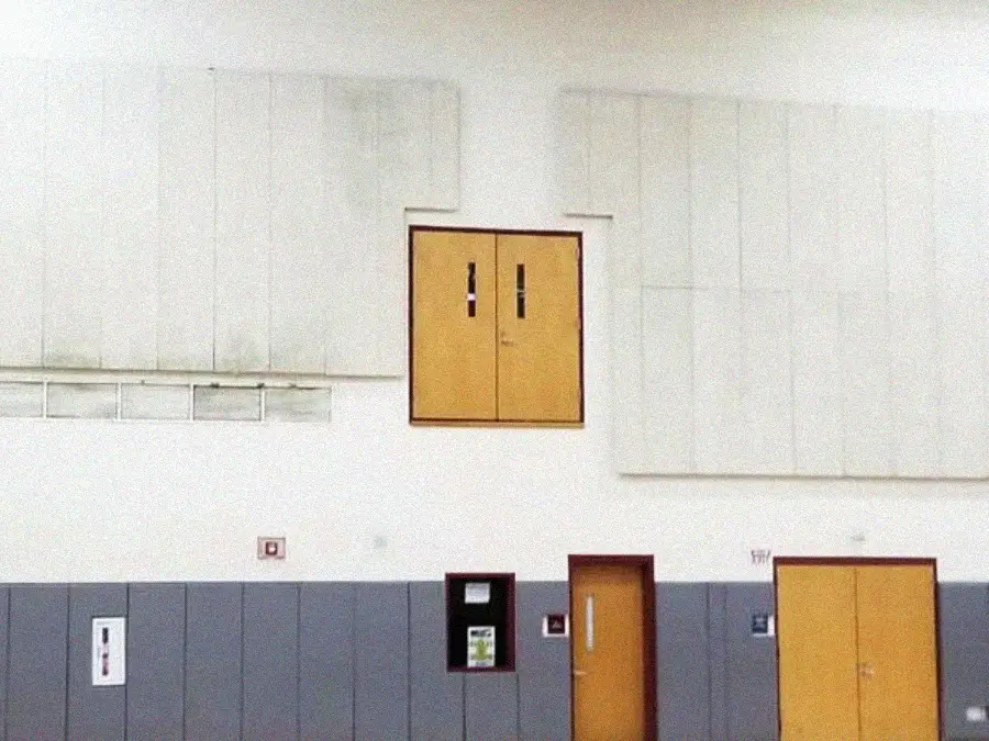 A gymnasium wall with two doors: one at floor level and another high above the ground, with no visible stairs or ladder leading to it. The wall is mostly white with gray lower panels.