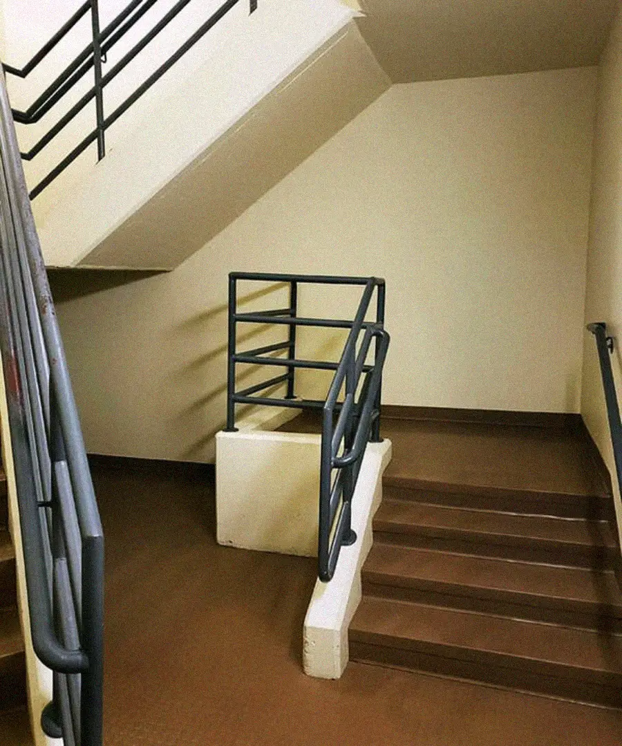 A staircase with brown steps and metal railings leads to a small landing, blocked by a railing, preventing further movement up or down. The stairs appear to go nowhere, ending at the wall.