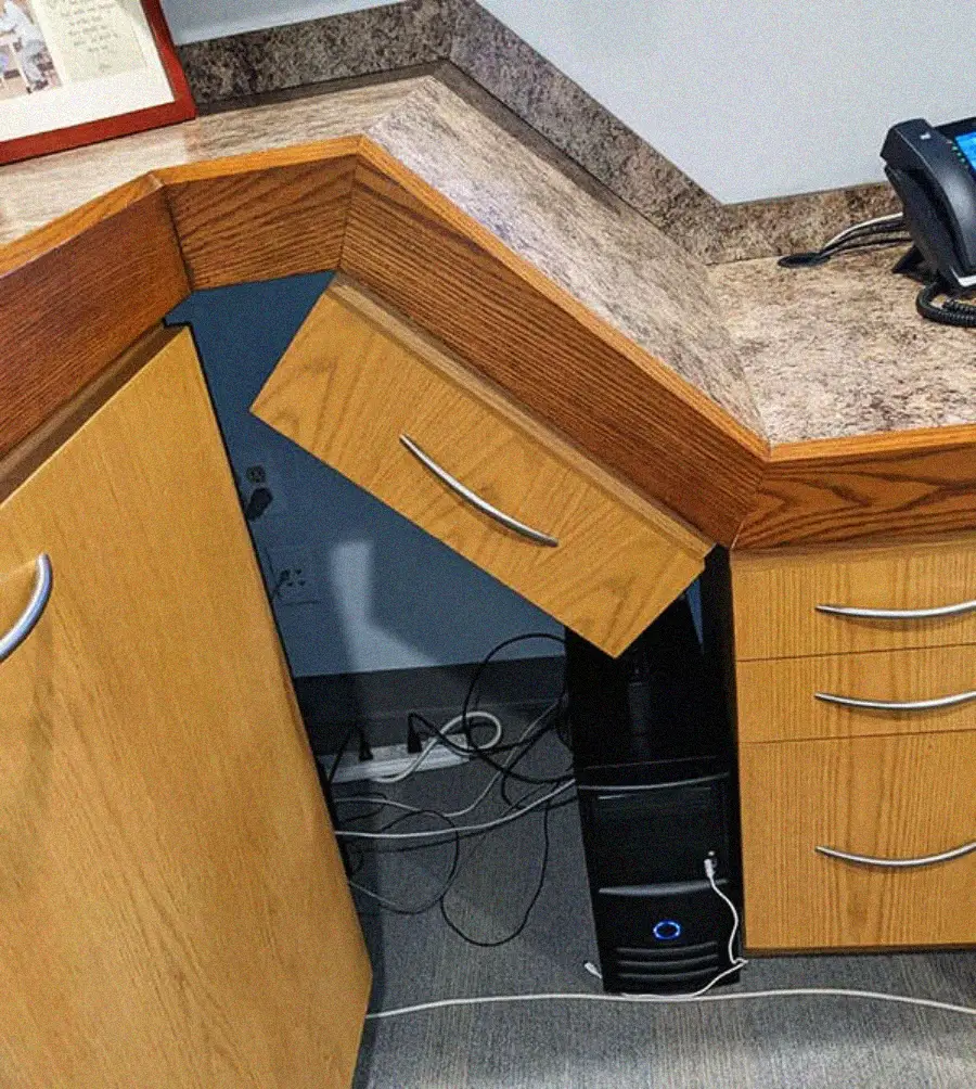 A drawer is open and blocked by the countertop, making it impossible to open fully due to poor design alignment with the desk corner. Nearby are a computer tower, cables, and a phone.