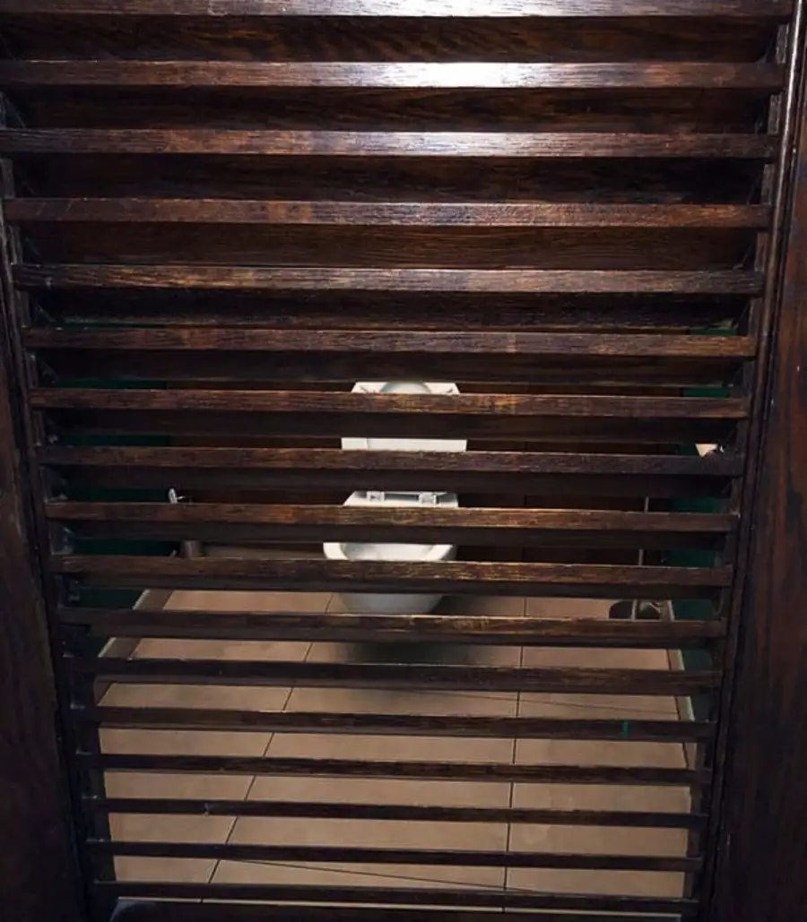 A toilet is visible behind dark wooden slats, revealing a bathroom with little privacy due to the gaps between the horizontal wooden bars.