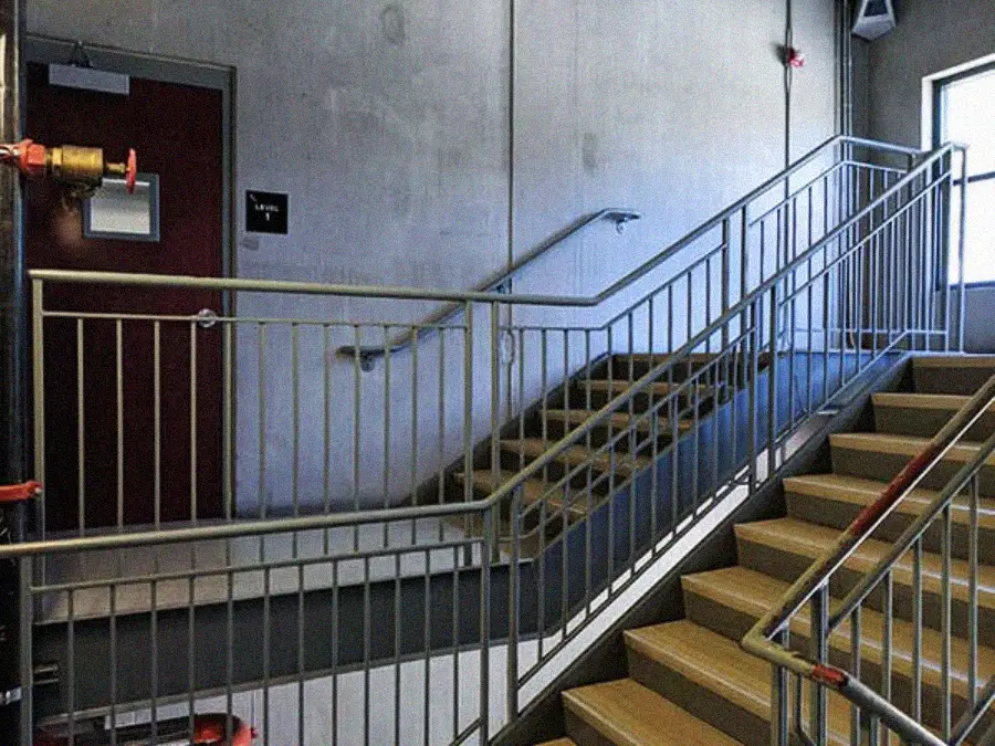 A stairwell with tan steps, metal railings, and gray walls. A closed red door is at the landing, and sunlight streams through a window on the upper right, casting shadows on the stairs.