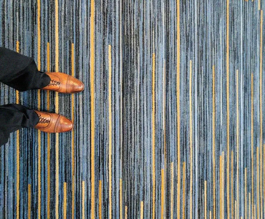 A person wearing brown dress shoes and black pants stands on a blue carpet with vertical yellow, blue, and gray stripes. Only the lower legs and feet are visible from a top-down view.