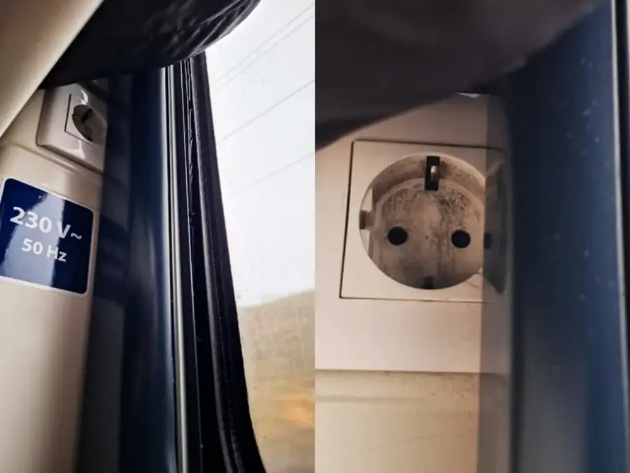 A European-style power outlet on a train is positioned next to a window, making it difficult to use standard plugs due to the narrow gap between the outlet and the window frame.