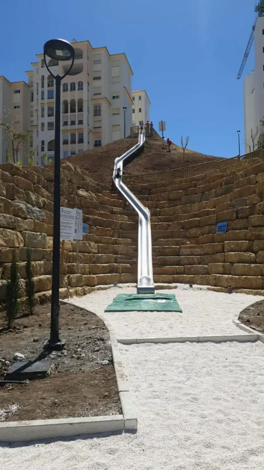 A long, silver playground slide descends a steep, terraced hill lined with stone walls, ending on a white gravel path in an urban area with tall buildings in the background.