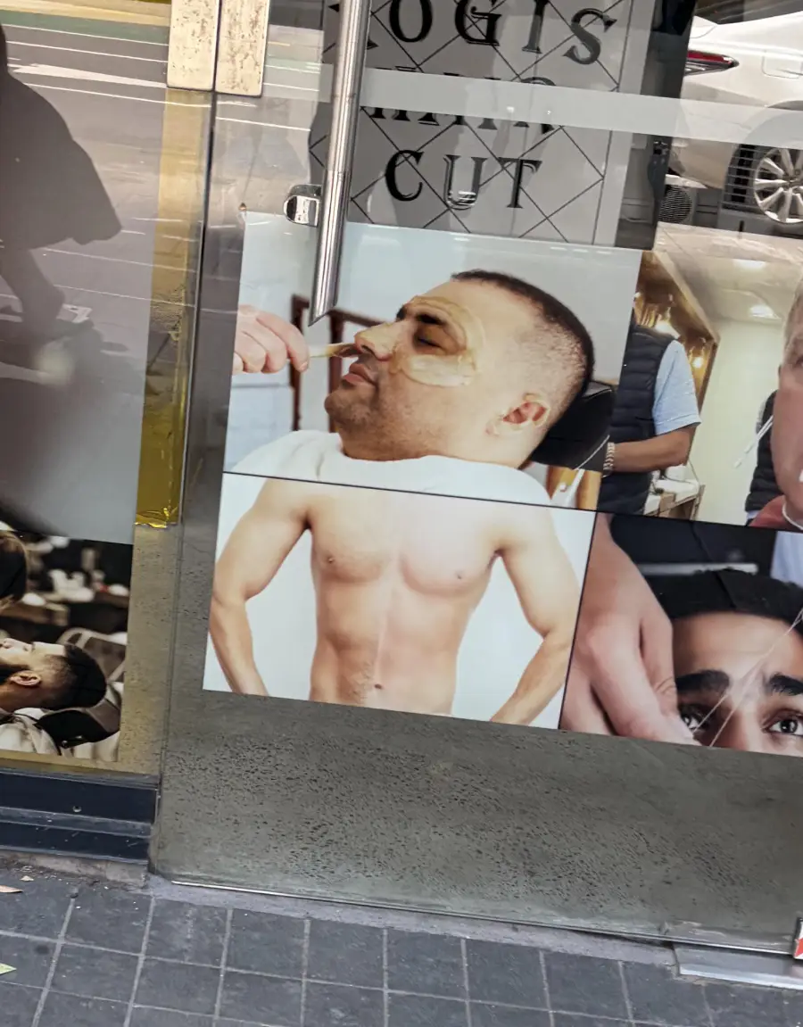 A storefront window displays photos of a man getting a nose wax, a shirtless man's torso, and close-ups of men's grooming treatments. Reflections of the street and a parked car are visible on the glass.