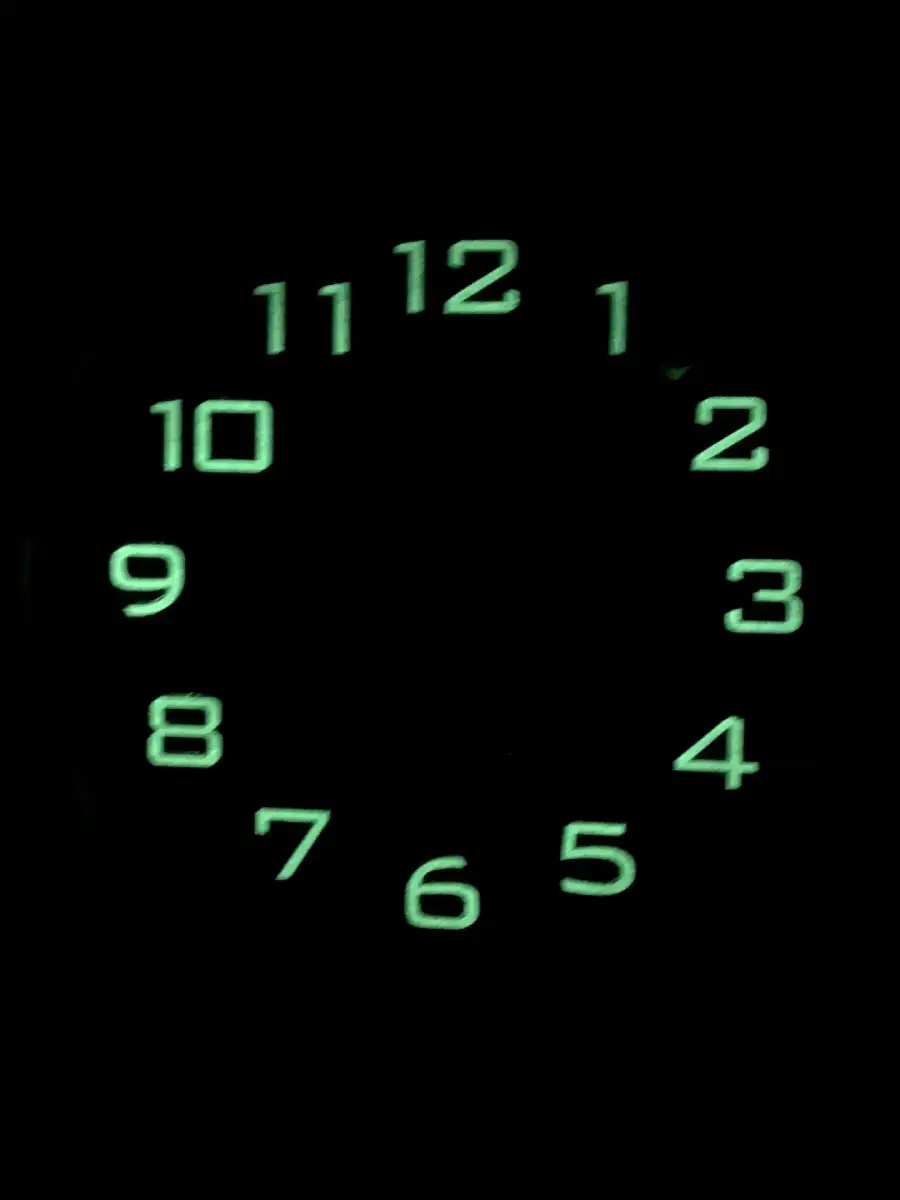Glow-in-the-dark clock face showing only the numbers 1 to 12 in a circle, illuminated in green against a black background, with no clock hands visible.