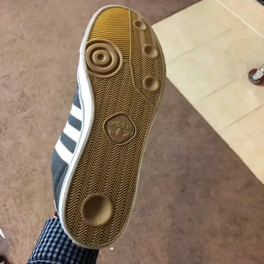 A close-up view of the sole of an Adidas shoe with a brown, textured grip featuring circular patterns and the Adidas logo, held up indoors over a carpeted and tiled floor.