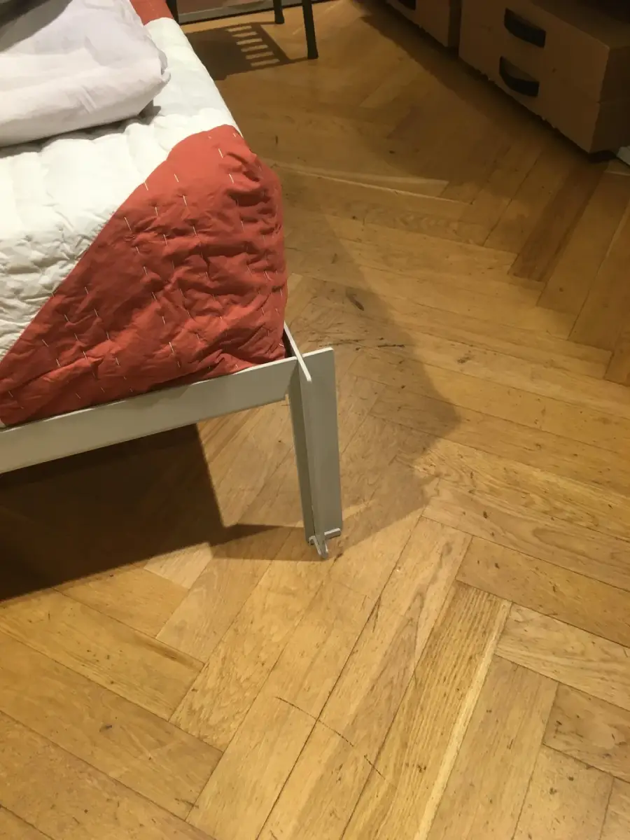 A metal bed frame leg misaligned with the floor stands at an angle, not touching the ground properly. The bed has a red and white quilt, and the room has wooden parquet flooring.
