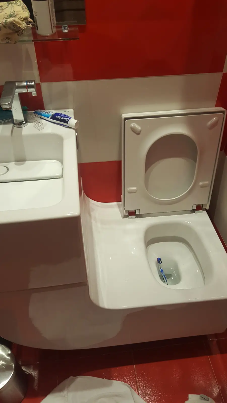 A toothbrush is lying inside a toilet next to a sink with toothpaste and a tube of cream on the edge, all against a red and white tiled wall.