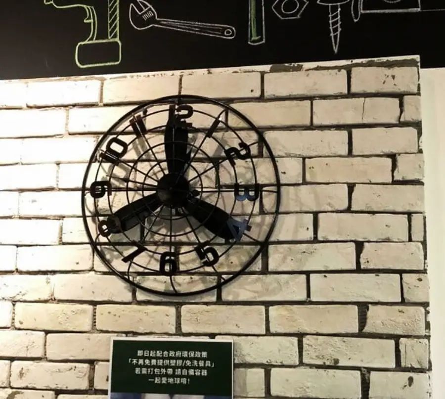 A wall clock with unusually large black hour and minute hands, making it difficult to see the time, is mounted on a white brick wall below a blackboard with green chalk drawings of tools.