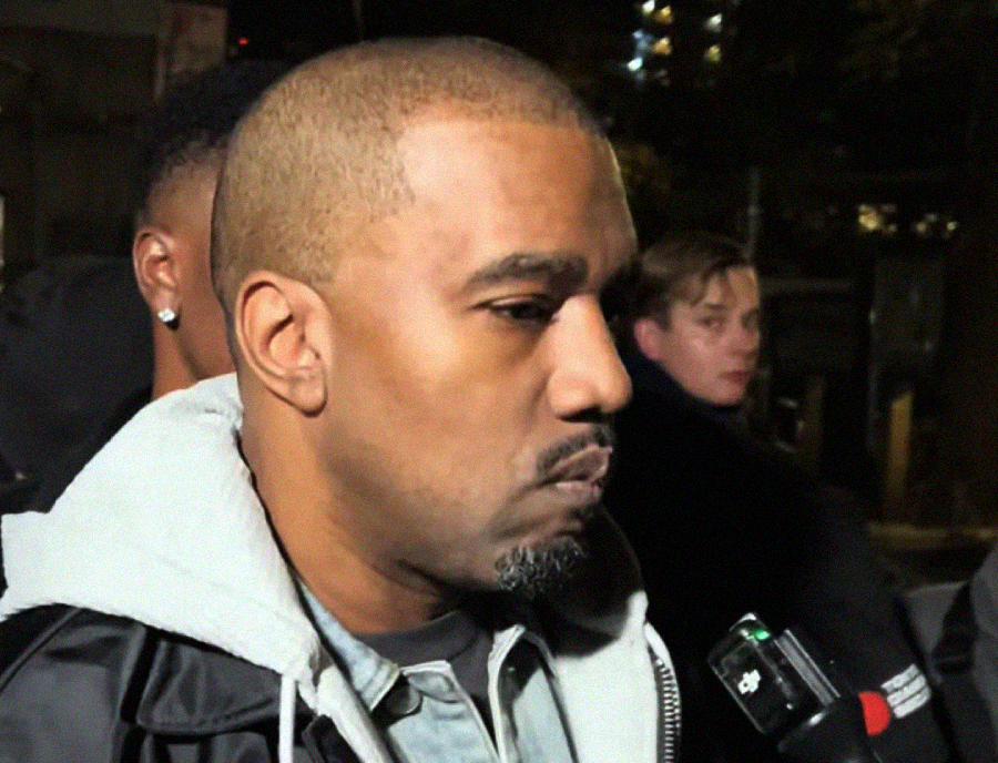 A man with a trimmed beard and shaved hairstyle wears a jacket with a hoodie underneath, standing outside at night and speaking into a microphone held by someone off camera. Other people are visible in the blurred background.