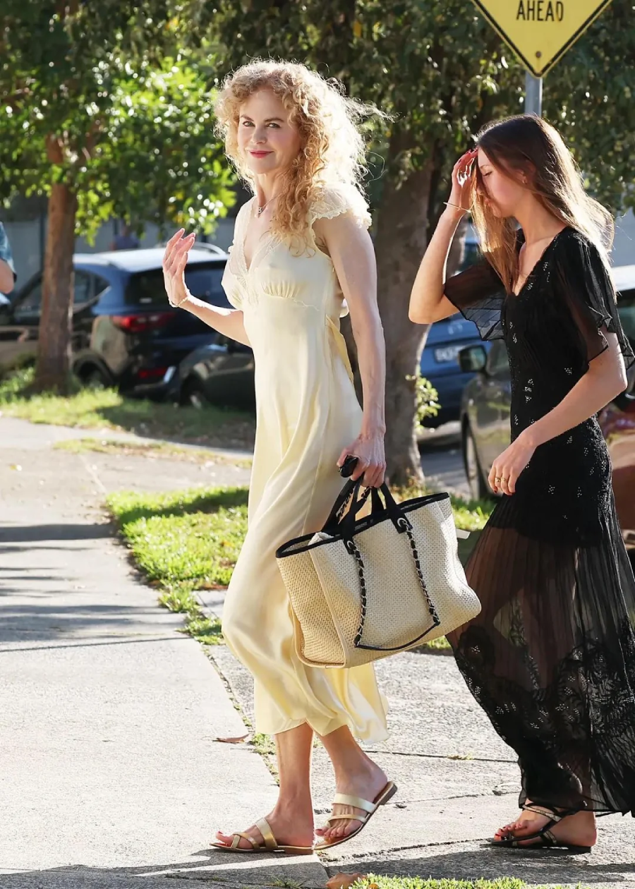 Two women walk on a sunny sidewalk. One wears a long, pale yellow dress and carries a woven bag, while the other wears a sheer, black dress and holds her hand to her forehead. Trees and parked cars are in the background.