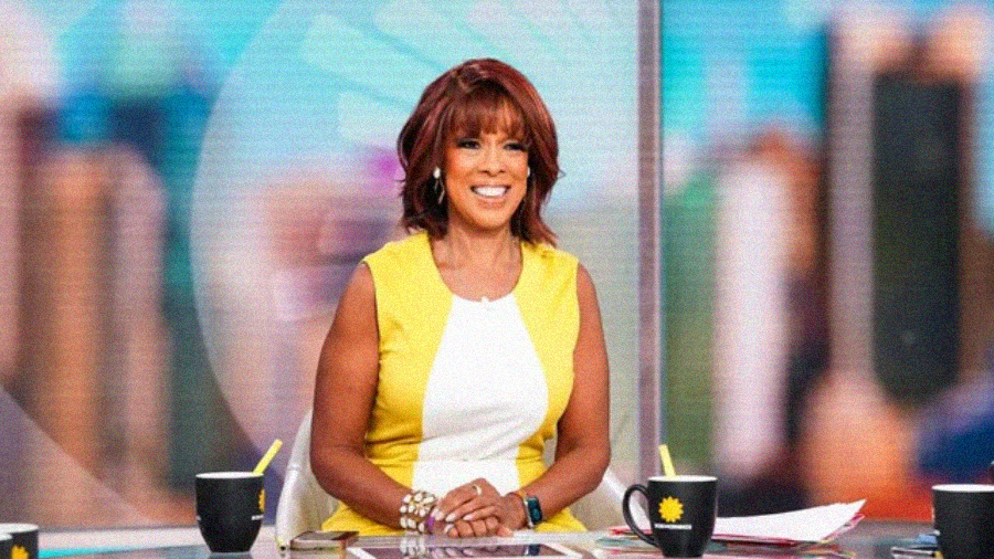 A smiling woman in a yellow and white dress sits at a news desk with two black mugs and papers in front of her. The blurred background features bright colors and a faint sunburst logo.