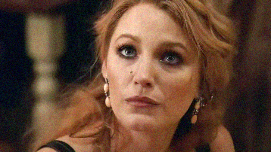 A woman with wavy reddish-blonde hair and dramatic makeup looks upward with a serious expression. She is wearing dangling earrings and a black outfit, with a blurred background behind her.