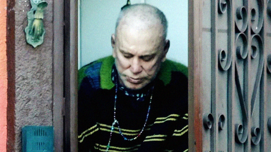 An older man with short gray hair and a striped sweater stands in a doorway, looking down, partially framed by a metal gate and stucco wall.