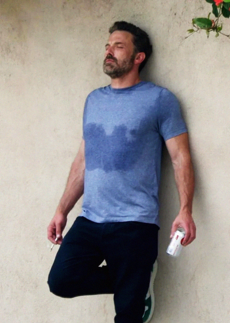 A man in a blue t-shirt with a sweat stain, dark pants, and green shoes leans against a beige wall. He holds a can in one hand and a cigarette in the other, looking upward with a tired expression.