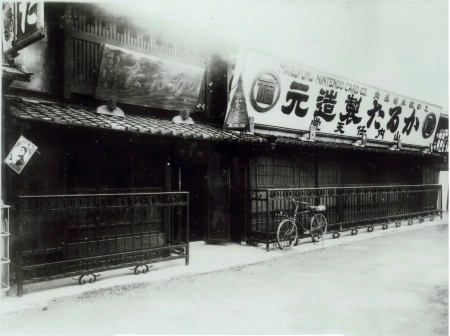 Black and white photo of an old Japanese building with a sign reading “Nintendo Playing Card Co.” in Japanese. A bicycle is parked outside, and the street in front appears empty.