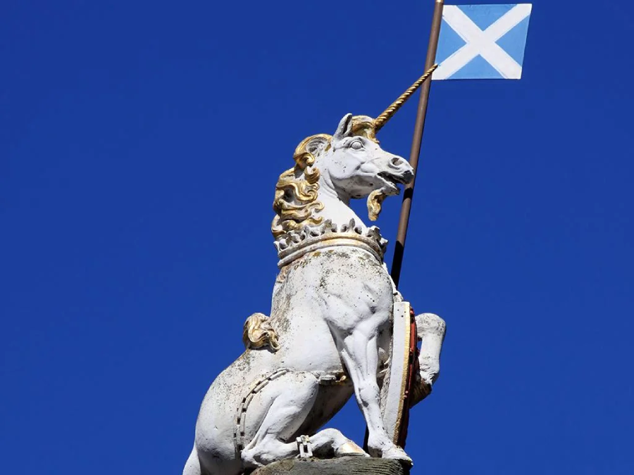 A white unicorn statue with a gold mane holds a Scottish flag against a clear blue sky.