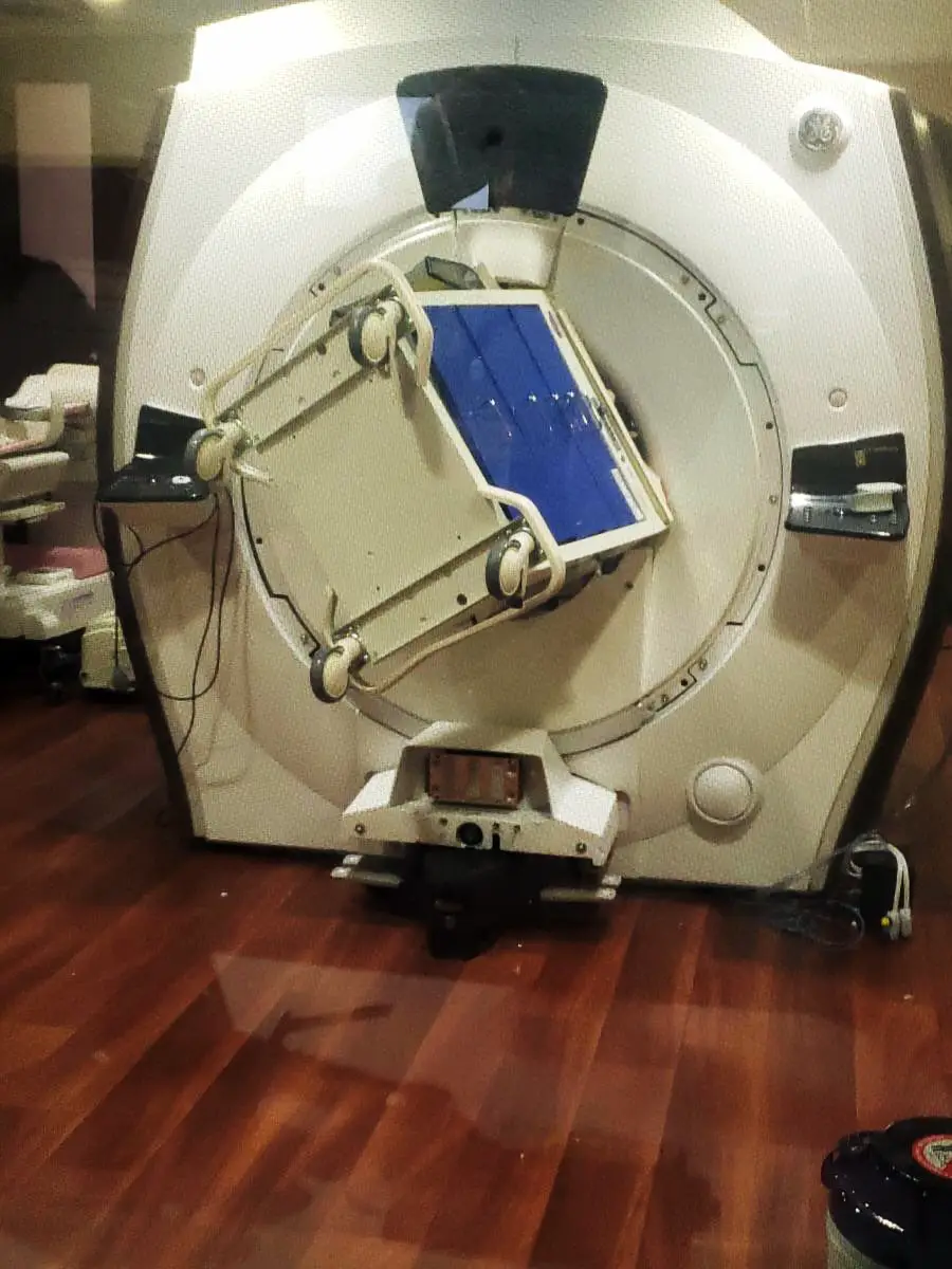 A large MRI machine with a chair lodged inside its circular opening sits on a wooden floor in a clinical room. The scene appears unusual and out of place for medical equipment.