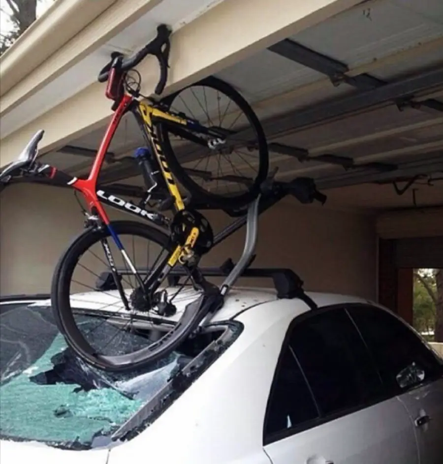 A road bike mounted on a car roof rack has struck the open garage door, causing the bike to bend backward and the car’s rear window to shatter.