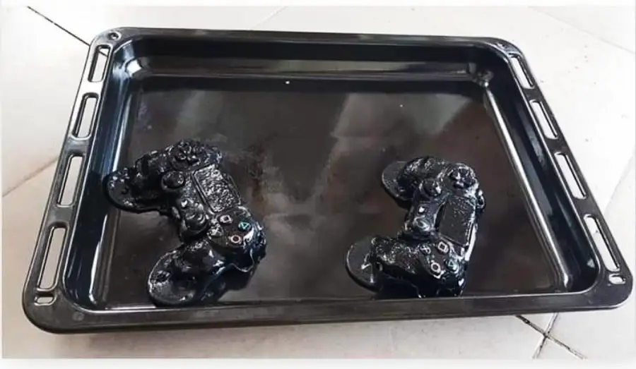 Two melted black video game controllers on a dark baking tray, placed on a tiled floor. The controllers appear heavily deformed from heat.