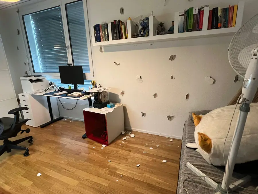 A home office with a desk, computer, fan, and chair is shown. The white wall is damaged with many holes and debris on the floor. A shelf with books and figures is above the desk; a plush toy and fan are on a gray sofa.