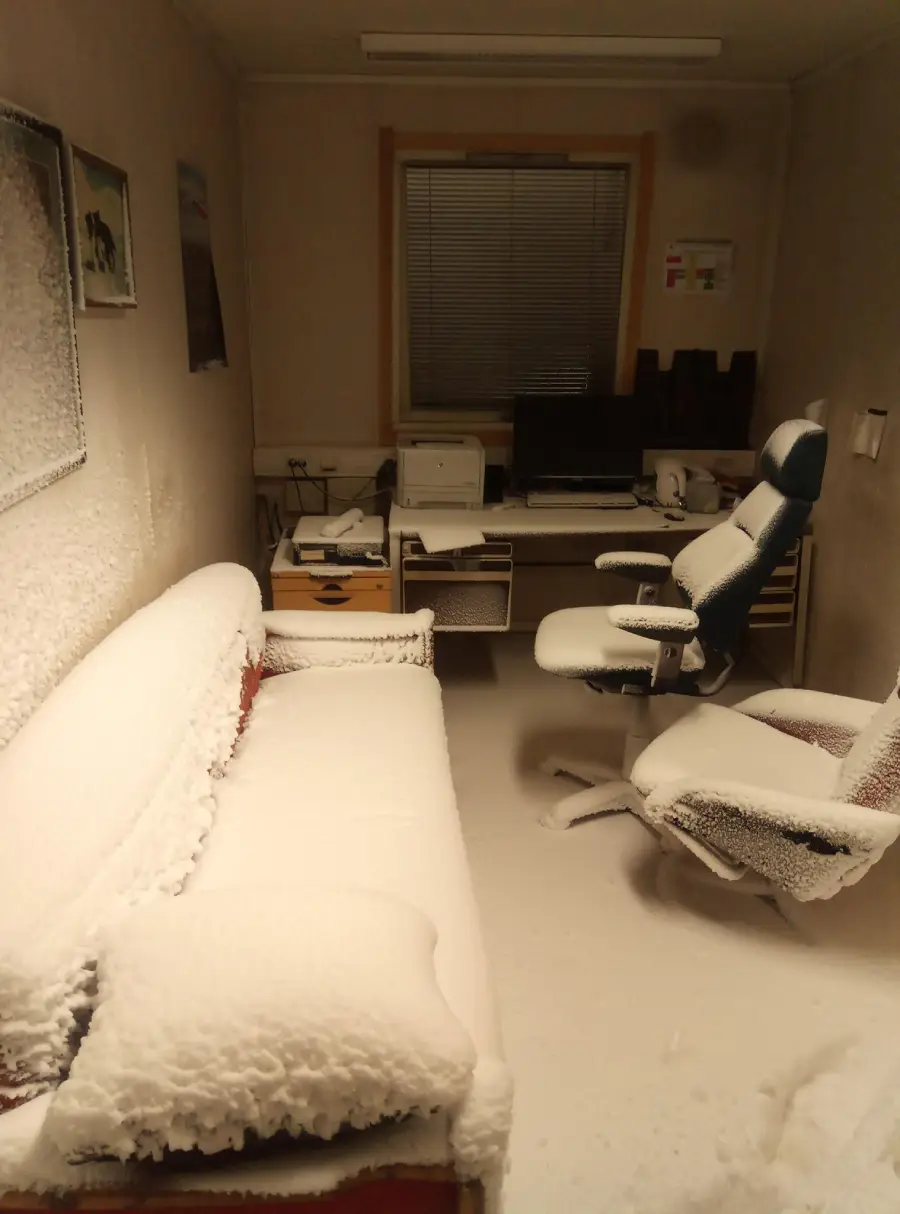 A small office with a desk, computer, office chair, and couch, all covered in a thick layer of snow, as if snow has blown in and settled throughout the room.