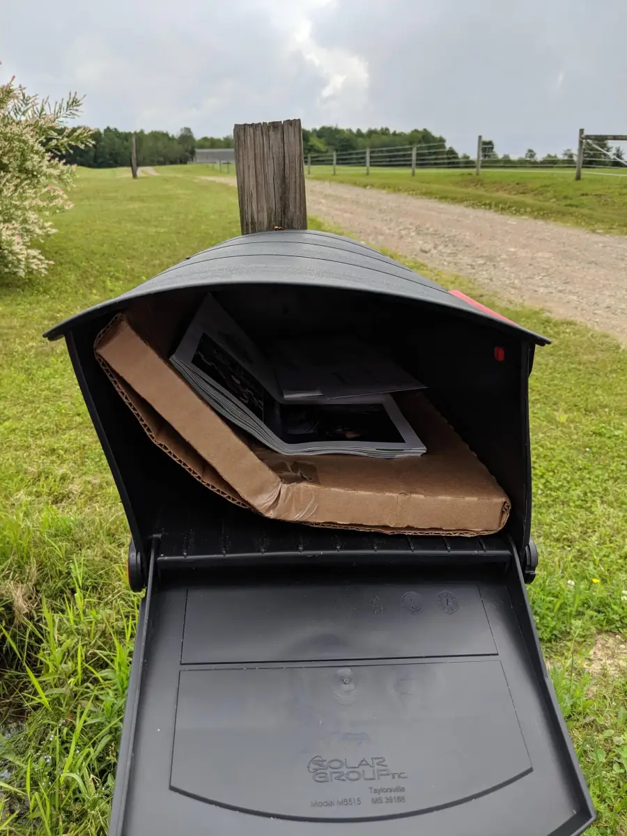 An open black mailbox contains a large, bent cardboard package and some letters, with a rural gravel road and grassy fields in the background under a cloudy sky.