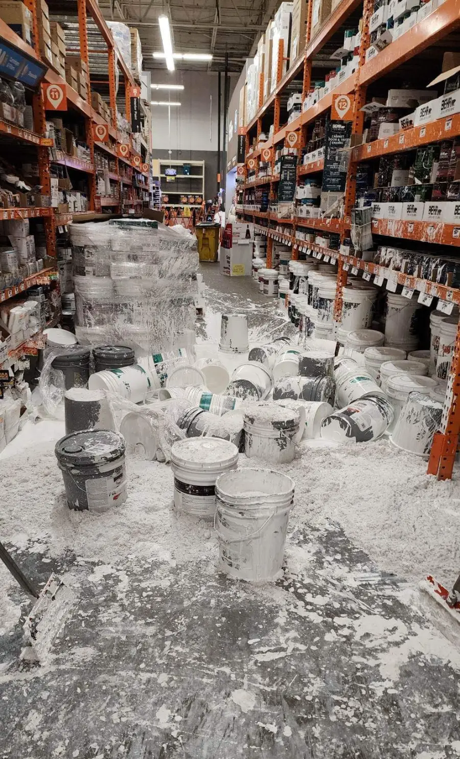 A hardware store aisle is covered in spilled white paint from multiple fallen buckets, creating a large mess on the floor among shelves stocked with paint cans.
