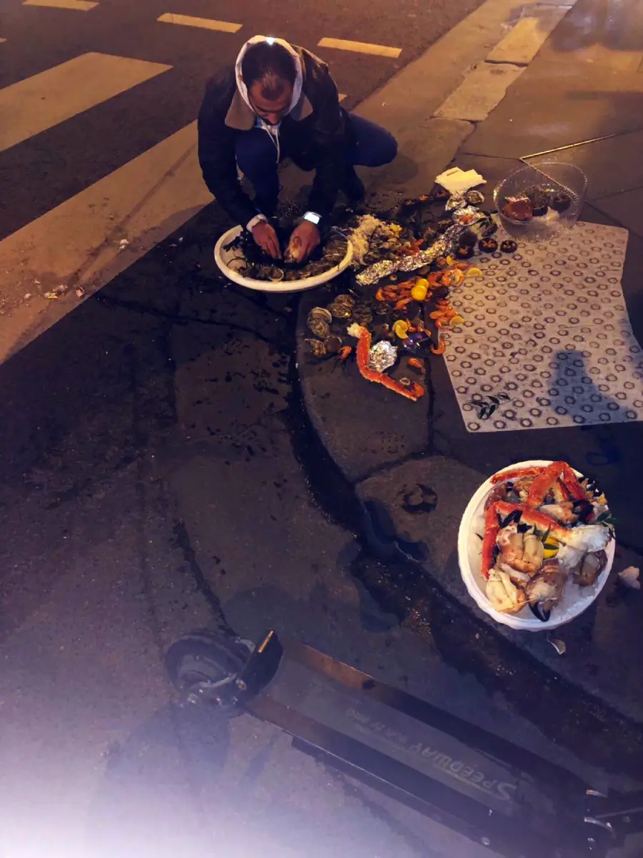A person crouches on a city sidewalk at night, sorting seafood—such as crabs and oysters—into large bowls. Various food items are scattered on a patterned mat nearby, next to a street curb and an electric scooter.