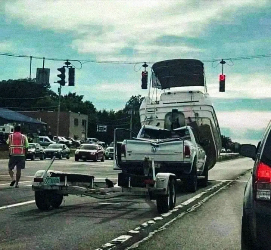 A white pickup truck towing a large boat on a trailer has tipped backward, lifting its front wheels off the road at a red light. A man in a safety vest stands nearby, and cars wait at the intersection.