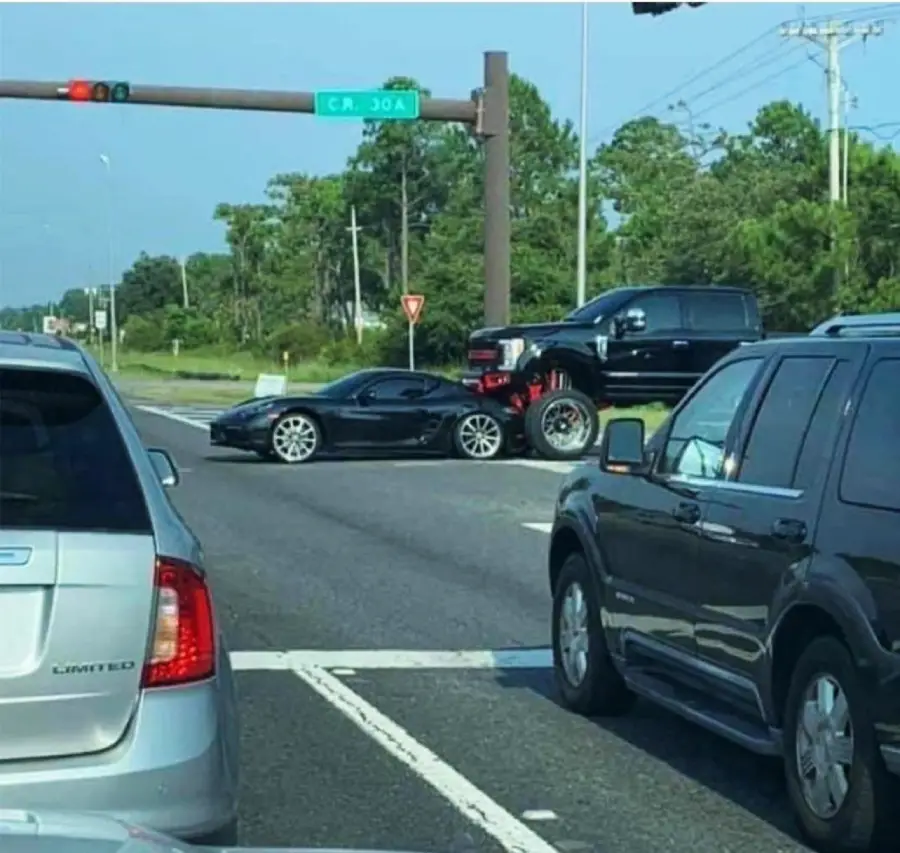 A lifted black pickup truck has driven on top of a black sports car in the middle of an intersection, while other vehicles wait at the traffic light. Trees and a street sign are visible in the background.