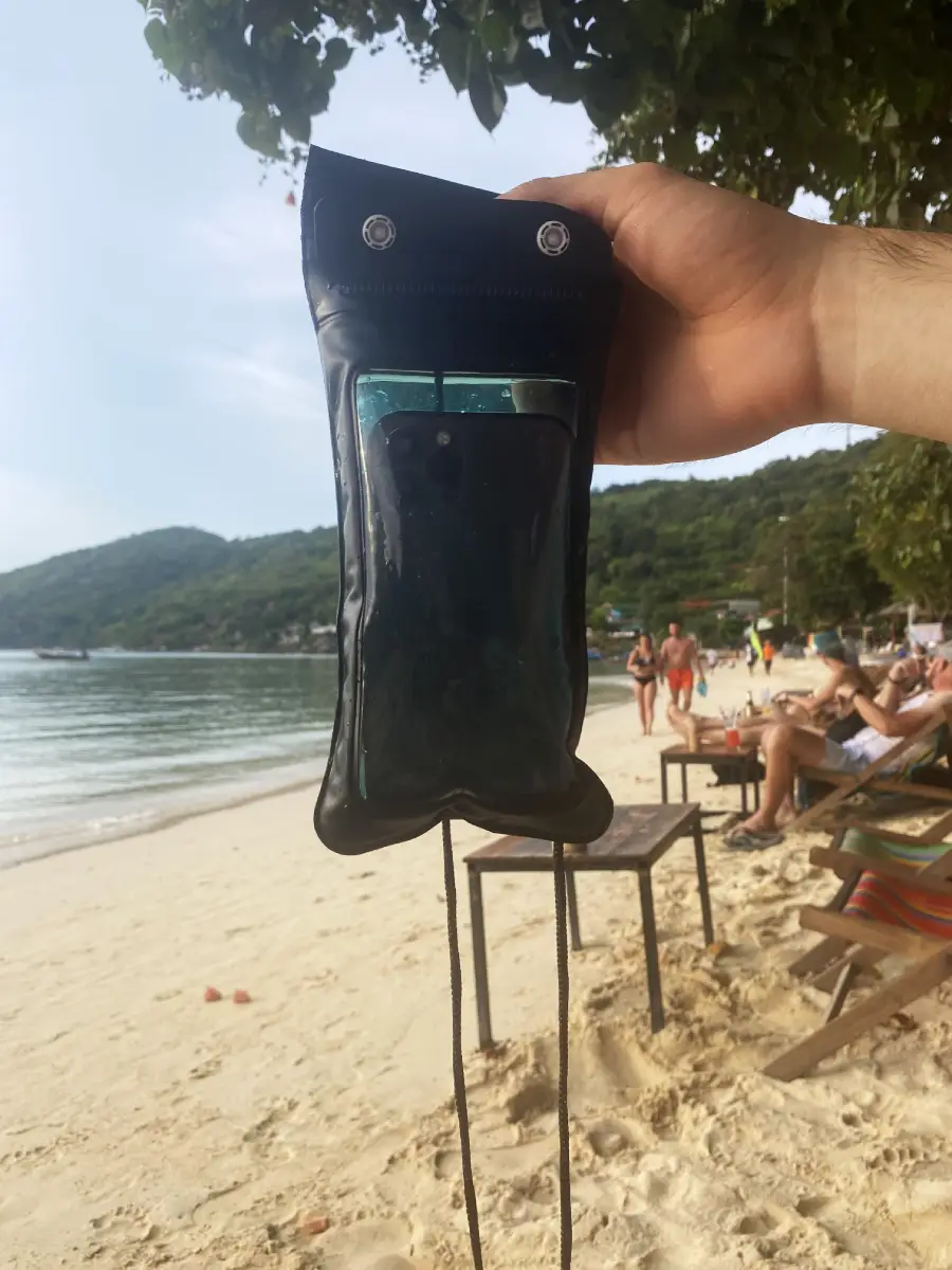 A hand holds a waterproof phone pouch containing a smartphone on a sandy beach, with people relaxing on lounge chairs, trees, and hills visible in the background.