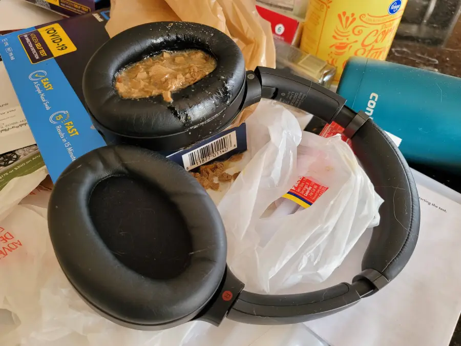 A pair of black over-ear headphones sits on a cluttered surface, with one earcup filled with spilled cereal and milk. Various containers, papers, and boxes surround the headphones.