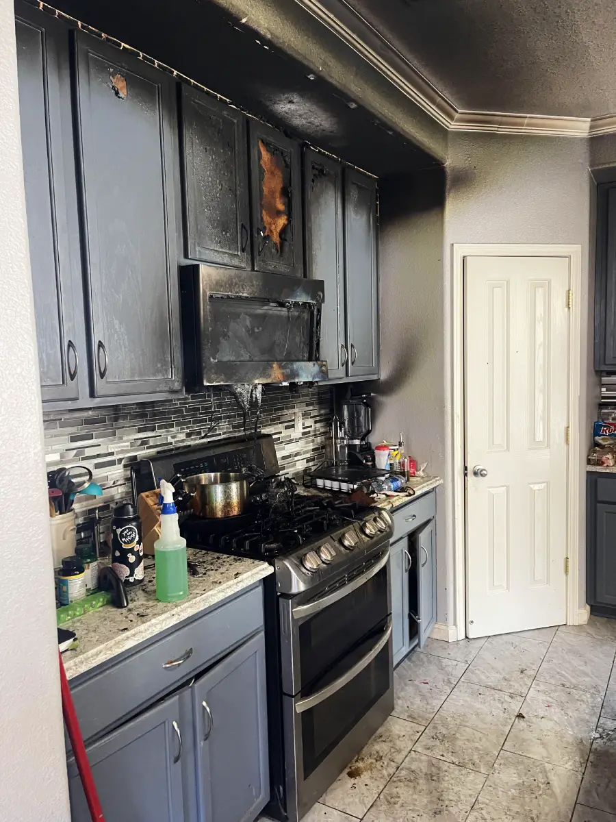 A kitchen with burned and soot-stained cabinets above the stove, showing fire damage. Pots and various kitchen items are on the counters, and the ceiling and walls are blackened from smoke.