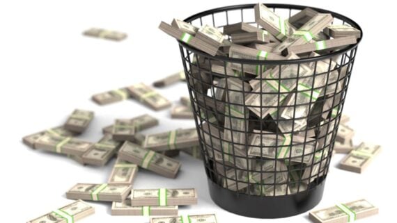 A black wire trash bin overflowing with bundles of U.S. dollar bills, with more bundles scattered on the white surface around the bin.