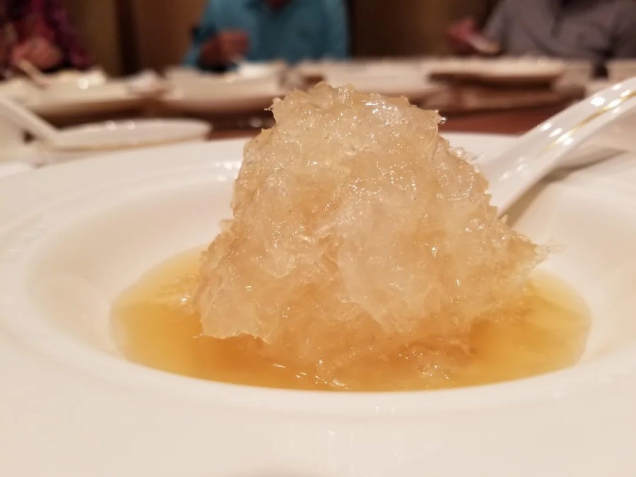 A close-up of a white plate with a mound of translucent, jelly-like food in a light brown broth. In the background, people are seated at a table with plates and utensils.