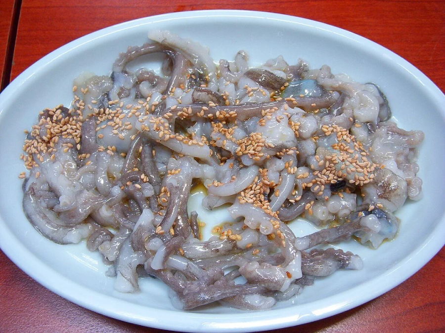 A white oval plate filled with sliced raw octopus, garnished with sesame seeds, on a reddish-brown wooden table.